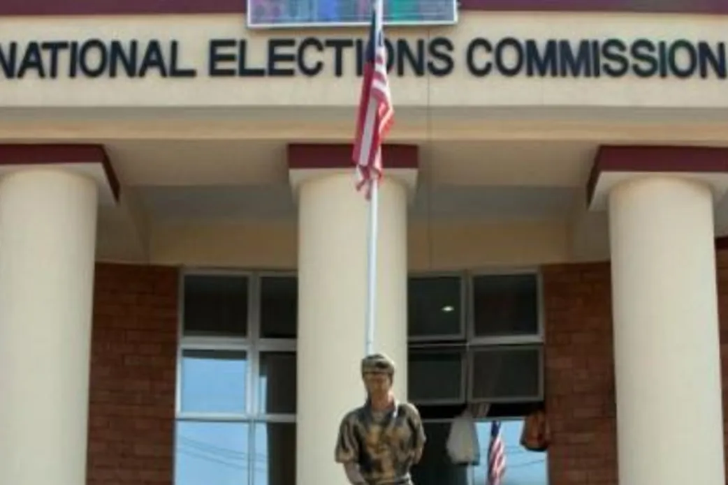 A peacekeeper standing in front of a flag pole flying the Liberian flag and in front of the Liberian National Elections Commission.